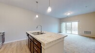 Located in Lakeville, MN, this well-appointed one-bedroom, two-bathroom unit offers a thoughtfully designed layout across a single floor. The open-concept kitchen and living area serves as the heart of the home, featuring a central kitchen island with a stone-look countertop, double stainless steel sink, and elegant pendant lighting overhead. Dark espresso cabinetry with brushed nickel hardware adds a refined contrast to the light hardwood-style flooring in the kitchen. Stainless steel appliances, including a refrigerator and range, complete the modern culinary space. The living room extends seamlessly from the kitchen, finished with plush neutral carpeting and a sliding glass door that opens to a private balcony with outdoor views of surrounding greenery. The generously sized bedroom offers large windows that flood the room with natural light and frame peaceful views of the tree line. A dedicated walk-in closet provides ample wire-rack storage. Both bathrooms are finished with rich dark vanity cabinetry, white countertops, and large mirrors, maintaining a consistent, cohesive aesthetic throughout the unit. The overall ambiance is clean, neutral, and move-in ready.