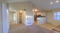 This single-floor residence showcases a thoughtfully designed open-concept layout that seamlessly connects the living area to the kitchen. Dramatic vaulted ceilings soar overhead in the main living space, creating an airy and expansive atmosphere that enhances the sense of openness throughout. The flooring transitions from soft, neutral carpeting in the living area to smooth hard-surface flooring in the kitchen, defining each zone while maintaining a cohesive aesthetic.

The kitchen is well-equipped with white cabinetry, dark countertops, stainless steel appliances including a refrigerator and microwave, and a peninsula that provides additional prep space and a natural divider between the cooking and living areas. Natural light filters into the space through windows, casting bright patches across the interior.

A standout feature of this residence is the generous built-in storage, highlighted by a large open shelving unit — ideal for organization and display. Additional doorways lead to further rooms, offering flexible use of the available space. Neutral tones throughout the walls, cabinetry, and flooring create a clean, versatile backdrop that suits a wide range of interior styles. The overall ambiance is bright, functional, and welcoming.