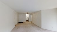 This single-floor unit presents a straightforward and functional layout centered around an open living area. The space is finished with light beige wall-to-wall carpeting that extends throughout the main living zone, contributing to a warm, neutral aesthetic. White walls and a white ceiling enhance the sense of openness and allow natural and artificial light to reflect evenly across the room.

A ceiling fan is centrally positioned in the living area, providing both air circulation and a modest lighting fixture. The front entry door is white and opens directly into the living space, with a dark floor mat marking the transition point at the threshold.

From the living area, there are clear sightlines into the kitchen, which features white cabinetry, light countertops, and what appears to be standard kitchen appliances. The kitchen area is distinguished by a tile or hard-surface floor, contrasting with the carpeted living zone. Additional doorways visible from the main space suggest access to further rooms within the unit.

Overall, the space presents a clean, minimalist environment with a simple color palette of whites and neutrals, offering a versatile and move-in ready interior suited to a variety of furnishing styles.