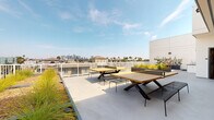 Located in the Koreatown neighborhood of Los Angeles, this rooftop amenity deck at 833 Fedora St offers a well-designed outdoor living experience with striking views of the downtown LA skyline. The rooftop is finished with a smooth concrete deck surface and is enclosed by a white metal railing that frames unobstructed sightlines toward the city and surrounding palm tree-lined neighborhood.

The space is divided into distinct functional zones. A social lounge area features a teak-framed sectional sofa with plush cushions centered around a sleek rectangular fire pit table, alongside a wall-mounted flat-screen television — ideal for outdoor gatherings day or night. Adjacent to this, a dining area provides multiple large wooden picnic-style tables with matching benches, accommodating groups of various sizes.

Integrated into the rooftop's design are generous raised planter beds finished in a dark charcoal tone, filled with ornamental grasses, shrubs, and drought-tolerant plantings that add texture and greenery to the hardscape. A partially covered corridor connects different sections of the deck, lined with additional planters featuring manicured boxwood hedges against a clean white exterior facade.

The overall aesthetic is modern and minimalist, combining natural wood elements with dark metal accents against a predominantly white architectural backdrop, creating a cohesive and sophisticated outdoor environment.