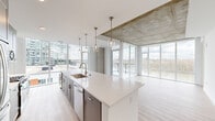 This well-appointed single-floor unit offers a thoughtfully designed open-concept layout that seamlessly integrates the kitchen, dining, and living areas into one cohesive space. The kitchen serves as a central showpiece, anchored by a generous white quartz waterfall island with an undermount sink and brushed gold faucet. A row of pendant lights hangs above, complementing the full suite of stainless steel appliances and grey shaker cabinetry that lines the walls. Exposed concrete ceiling elements add an industrial character that contrasts elegantly with the crisp white finishes throughout.

Floor-to-ceiling windows dominate the living area, flooding the space with natural light and framing expansive views of a nearby river, bridge, and urban skyline. A glass-paneled balcony door extends the living space outdoors. Light wood-tone flooring flows continuously throughout the unit, enhancing its airy, modern feel.

The bedroom features large windows with city views and an exposed concrete ceiling, maintaining the industrial-modern aesthetic. Two closets provide ample storage, including a well-organized walk-in. Both bathrooms are finished in clean, neutral tones; one includes a tub-shower combination, a slate-blue vanity with a white countertop, and a window with natural light.