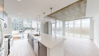 This well-appointed single-floor unit offers a thoughtfully designed open-concept layout that seamlessly integrates the kitchen, dining, and living areas into one cohesive space. The kitchen serves as a central showpiece, anchored by a generous white quartz waterfall island with an undermount sink and brushed gold faucet. A row of pendant lights hangs above, complementing the full suite of stainless steel appliances and grey shaker cabinetry that lines the walls. Exposed concrete ceiling elements add an industrial character that contrasts elegantly with the crisp white finishes throughout.

Floor-to-ceiling windows dominate the living area, flooding the space with natural light and framing expansive views of a nearby river, bridge, and urban skyline. A glass-paneled balcony door extends the living space outdoors. Light wood-tone flooring flows continuously throughout the unit, enhancing its airy, modern feel.

The bedroom features large windows with city views and an exposed concrete ceiling, maintaining the industrial-modern aesthetic. Two closets provide ample storage, including a well-organized walk-in. Both bathrooms are finished in clean, neutral tones; one includes a tub-shower combination, a slate-blue vanity with a white countertop, and a window with natural light.