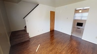 This two-floor home offers a practical and well-organized layout across its living spaces. The main level features a welcoming living area with rich, dark hardwood-style flooring that reflects natural light streaming in through the windows. A staircase with carpeted treads leads to the upper floor, while a separate entry area provides a bright and airy introduction to the home, also finished with the same warm hardwood-style flooring.

The kitchen is equipped with warm wood cabinetry spanning both upper and lower sections, offering generous storage throughout. A double-basin sink, laminate countertops, a white electric range, and a full-size refrigerator complete the functional cooking space. The kitchen also features tile flooring, a ceiling fan, and direct access to an exterior door.

The bedrooms are located on the upper floor and are finished with plush carpeting. Each bedroom includes a dedicated closet with a sliding wood-panel door and a clothing rod, providing ample storage. Natural light filters in through the windows, creating a comfortable atmosphere.

The bathroom features classic white subway-style wall tiling, a pedestal sink, a mirrored medicine cabinet with overhead lighting, and a bathtub with an overhead shower — offering a clean, timeless design throughout.