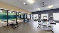Located at Jefferson Commons in Kalamazoo, Michigan, this single-story recreational facility offers a comprehensive fitness and sports experience. The fitness center is generously sized, featuring a wide array of strength training equipment including cable machines, multi-station weight systems, and free weights, complemented by an expansive floor-to-ceiling mirror wall that enhances the sense of space. The cardio zone is well-appointed with treadmills, elliptical trainers, and stair climbers, all positioned beneath ceiling fans and recessed lighting. Large windows and French doors along the perimeter allow abundant natural light to fill the workout area, creating a bright and energizing environment. A connecting corridor features hardwood-style flooring, a water fountain, vending machines, and additional strength equipment, providing a practical transition between zones. The highlight of the facility is the full-size indoor basketball court, boasting a polished hardwood floor, a regulation backboard and hoop, padded wall panels for player safety, and high clerestory windows that introduce natural light from above. The court's soaring ceiling height and open layout make it suitable for a variety of recreational activities beyond basketball, offering a versatile space for active use year-round.
