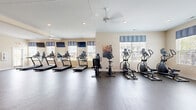 Located at 915 Galloway Rd in Galloway, Ohio, this fitness center offers a comprehensive, single-floor workout facility designed to support a wide range of exercise routines. The space is thoughtfully laid out to maximize functionality, with distinct zones dedicated to cardio training, strength equipment, and open-floor exercise.

The cardio area features a row of treadmills and elliptical machines positioned along mirrored walls and large windows dressed with stylish valances, allowing natural light to fill the room while offering views of the surrounding neighborhood. Wall-mounted televisions provide entertainment during workouts. Ceiling fans ensure comfortable air circulation throughout.

The strength training section is equipped with a leg press machine, a cable/weight station, a dumbbell rack, and a workout bench, offering versatile options for resistance training. Colorful exercise balls and additional accessories are available for functional fitness and stretching routines.

The open exercise area features durable rubberized flooring, providing a safe and spacious surface for floor exercises, stretching, or group activities. Full-length mirrors along the walls enhance the sense of space and assist with form during workouts.

The interior maintains a clean, neutral aesthetic with light-toned walls, recessed lighting, and a bright, welcoming ambiance that makes the fitness center an inviting space for daily use.