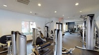 This fitness center is designed to support a comprehensive workout experience within a single-floor layout. The space is divided into distinct workout zones, with strength training equipment positioned toward the front near a large window that allows natural light to fill the area. Multiple cable and weight machines, appearing to be from a recognized commercial brand, are arranged to allow comfortable circulation between stations.

Recessed lighting throughout the textured white ceiling ensures consistent illumination across the entire facility. The flooring features a dark, rubberized surface in the strength training area, providing both durability and safety underfoot. Toward the rear of the space, a separate zone houses cardio equipment, including treadmills and other aerobic machines, set against a contrasting deep blue-grey accent wall that adds visual depth to the room.

A large mirrored wall in the cardio section enhances the sense of space while allowing users to monitor their form. Access doors with glass panels connect the different areas, contributing to an open, airy feel. The overall design is functional and modern, with a clean, neutral color palette that creates a focused and energizing environment suitable for a range of fitness activities.