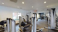 This fitness center is designed to support a comprehensive workout experience within a single-floor layout. The space is divided into distinct workout zones, with strength training equipment positioned toward the front near a large window that allows natural light to fill the area. Multiple cable and weight machines, appearing to be from a recognized commercial brand, are arranged to allow comfortable circulation between stations.

Recessed lighting throughout the textured white ceiling ensures consistent illumination across the entire facility. The flooring features a dark, rubberized surface in the strength training area, providing both durability and safety underfoot. Toward the rear of the space, a separate zone houses cardio equipment, including treadmills and other aerobic machines, set against a contrasting deep blue-grey accent wall that adds visual depth to the room.

A large mirrored wall in the cardio section enhances the sense of space while allowing users to monitor their form. Access doors with glass panels connect the different areas, contributing to an open, airy feel. The overall design is functional and modern, with a clean, neutral color palette that creates a focused and energizing environment suitable for a range of fitness activities.