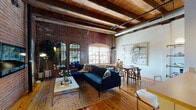 This single-floor loft showcases a compelling blend of historic industrial character and contemporary design. The centerpiece of the space is the expansive open-plan living and dining area, defined by soaring exposed wood beam ceilings with visible ductwork, warm hardwood floors, and dramatic full-height exposed brick walls. Arched brick window surrounds frame natural light that flows generously into the living space, highlighting the rich textures throughout. The living area accommodates a comfortable seating arrangement anchored by a patterned area rug, while the dining zone features a solid wood table paired with classic Wishbone chairs and is accented by large-scale artwork.

The kitchen is efficiently designed with white cabinetry, a galley-style layout, and a peninsula island, benefiting from the same soaring ceilings visible throughout the main living areas. Two bedrooms offer a quieter retreat, with neutral carpet, clean white walls, and understated lighting that contrast the industrial energy of the main living space. The bathroom is finished with light tile, a full tub-shower combination, and simple, clean fixtures. Throughout the unit, the interplay between raw, original architectural elements and modern furnishings creates a warm yet dynamic industrial ambiance.