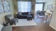 This single-floor residence offers a thoughtfully designed open-concept layout that seamlessly connects the living and dining areas, creating a spacious and inviting atmosphere throughout. Rich hardwood flooring runs continuously across the space, lending warmth and a cohesive feel to the interior.

The living area is anchored by a contemporary dark gray sectional sofa accented with bold geometric throw pillows, complemented by a matching accent chair and a soft cream area rug. Large windows dressed in elegant patterned curtains flood the room with natural light, highlighting the neutral wall tones and carefully curated wall décor, including modern metal art pieces and decorative frame arrangements.

The adjacent dining area flows naturally from the living space and features a round dining table surrounded by upholstered chairs, set atop a coordinating area rug. A track lighting fixture overhead provides focused illumination, while additional windows continue to bring in bright, natural light.

The overall ambiance is modern yet comfortable, with a consistent color palette of grays, whites, and warm neutrals that ties the spaces together. The open layout and generous flow between rooms make this home feel both functional and welcoming, ideal for everyday living and entertaining alike.