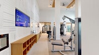 Located at 2001 E Fourth Street in Fort Worth, Texas, this fitness center offers a thoughtfully designed workout facility that blends functionality with upscale aesthetics. The single-floor space is generously proportioned, providing ample room for a comprehensive range of exercise equipment and activities.

The facility is characterized by soaring ceilings with warm wood beam accents and a clean, bright color palette of whites and soft neutrals. Multiple zones are defined throughout the space, including a strength training area, a dedicated free weights section, and a cardio zone. The free weights area features a striking slatted wood ceiling with integrated linear lighting and floor-to-ceiling mirrors that enhance the sense of depth.

Premium Matrix-brand equipment is distributed throughout, including cable machines, resistance stations, treadmills, stair climbers, and stationary bikes. The cardio zone is anchored by expansive floor-to-ceiling windows that flood the space with natural light and offer open views of the surrounding outdoor landscape.

Along one wall, a built-in wood storage unit and workstation with mounted flat-screen TVs serve as a functional amenity hub. A bold Fort Worth-themed mural decorates the entry wall, adding a distinctive local character to the space. The overall ambiance is polished, modern, and inviting, creating an environment well-suited for a focused and energizing workout experience.