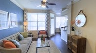 This single-floor residence features a well-appointed living area defined by rich, dark hardwood flooring and elegant crown molding that frames the space with a classic architectural touch. The living room is generously lit by multiple large windows adorned with patterned curtains and horizontal blinds, allowing abundant natural light to fill the interior. A ceiling fan with an integrated light fixture serves as a functional centerpiece overhead, contributing to the room's comfortable ambiance.

The living area is thoughtfully arranged with a neutral-toned sofa, an accent chair with warm orange cushioning, and a sleek glass-top coffee table resting on a plush area rug. A half-wall partition with a pass-through opening connects the living room to an adjoining dining area, which is visible in the background and features a chandelier and bar-style storage. Decorative plants and floral arrangements are placed throughout, adding organic warmth to the space. The combination of crown molding, hardwood floors, and layered lighting gives the home a polished, inviting character throughout.