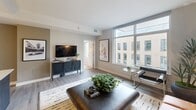 Located in McLean, VA, this thoughtfully designed two-bedroom, one-bathroom apartment offers a modern aesthetic with high-quality finishes throughout. The open-concept layout maximizes the use of space, creating a seamless flow between the living, dining, and kitchen areas. Large floor-to-ceiling windows flood the living room with natural light while offering views of the surrounding cityscape, complementing the light hardwood flooring and contemporary furnishings.

The kitchen is a standout feature, boasting dark cabinetry, a subway tile backsplash, stainless steel appliances, and a generous quartz-topped island with an integrated sink and seating — ideal for casual dining or entertaining. Pendant lighting adds a refined touch overhead.

The primary bedroom is a well-proportioned retreat, featuring a mirrored accent wall, built-in lighting at the headboard, and access to a closet. The second bedroom offers a cozy and versatile space with a closet, neutral tones, and a playful wall mural.

The bathroom is clean and modern, featuring a double-sink vanity with dark wood cabinetry, quartz countertops, a glass-enclosed walk-in shower, and elegant tile work throughout. The overall ambiance of this apartment is contemporary, polished, and move-in ready.