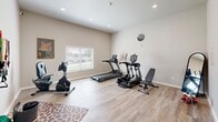 Located in Lawton, Oklahoma, this dedicated exercise room offers a compact yet efficiently organized fitness space designed to support a full range of workout routines. The room features light wood-look flooring throughout, neutral-toned walls, and recessed lighting that creates a bright and energizing atmosphere. A window fitted with blinds allows natural light to filter in while maintaining privacy, and a wall-mounted mini-split unit ensures comfortable climate control year-round.

The space is equipped with a thoughtful selection of cardio and strength training equipment, including a treadmill, elliptical trainer, and stationary bike — all from quality brands. A dedicated resistance training area features a wall-mounted pegboard displaying an organized array of resistance bands and suspension training tools, alongside a workout bench, balance board, and free weights. A full-length mirror supports form-checking during exercise routines.

French-style glass doors connect the exercise room to an adjoining area, lending an open and airy feel. Motivational posters and a wall clock add functional and decorative touches, while a patterned area rug grounds the space. This well-appointed fitness room provides everything needed for a comprehensive workout in a clean, purpose-built environment.