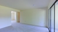 This single-floor space presents a straightforward and functional layout designed to accommodate a variety of uses. The interior is finished with soft, neutral wall tones in a warm off-white palette, creating a bright and welcoming atmosphere throughout. The flooring features wall-to-wall carpeting in a light gray hue, adding a comfortable underfoot feel and contributing to the overall understated aesthetic. Ceiling-mounted HVAC vents are integrated cleanly into the white ceilings, ensuring efficient climate control without disrupting the visual flow of the space. An open doorway connects the main room to an adjacent area, suggesting a multi-room layout that allows for easy movement between spaces. The walls are smooth and clean, with standard electrical outlets positioned conveniently at baseboard level. The absence of furniture and décor highlights the generous proportions of the rooms, offering a blank canvas that can be adapted to suit a wide range of needs. Overall, the space delivers a clean, move-in-ready environment with a calm, neutral character that serves as an excellent foundation for any interior arrangement.