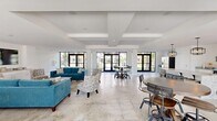 This single-floor space offers a generous, open-concept layout that seamlessly integrates the living room, dining area, and kitchen into one cohesive environment. The interior is defined by a clean, modern aesthetic with crisp white walls, coffered ceilings with structural beam detailing, and light polished stone tile flooring throughout that enhances the sense of spaciousness.

The living area is anchored by teal upholstered seating, a wood-top coffee table, and a wall-mounted flat-screen TV set above built-in white cabinetry with granite countertops. Natural light floods the space through an abundance of black-framed floor-to-ceiling windows and glass sliding doors, offering unobstructed views of the surrounding greenery and outdoor areas.

The dining zone features a round table positioned near a set of glass French doors, reinforcing the indoor-outdoor connection. The kitchen is equipped with a substantial granite-topped island, white shaker-style cabinetry, stainless steel appliances including a wine cooler and wall oven, and pendant lighting with an industrial-modern finish.

The entry corridor showcases full-height glazing and automatic glass doors, creating a welcoming, light-filled approach. Throughout, the architecture maintains a minimalist character, with the open layout, abundant natural light, and outdoor views contributing to an airy, contemporary ambiance.