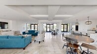 This single-floor space offers a generous, open-concept layout that seamlessly integrates the living room, dining area, and kitchen into one cohesive environment. The interior is defined by a clean, modern aesthetic with crisp white walls, coffered ceilings with structural beam detailing, and light polished stone tile flooring throughout that enhances the sense of spaciousness.

The living area is anchored by teal upholstered seating, a wood-top coffee table, and a wall-mounted flat-screen TV set above built-in white cabinetry with granite countertops. Natural light floods the space through an abundance of black-framed floor-to-ceiling windows and glass sliding doors, offering unobstructed views of the surrounding greenery and outdoor areas.

The dining zone features a round table positioned near a set of glass French doors, reinforcing the indoor-outdoor connection. The kitchen is equipped with a substantial granite-topped island, white shaker-style cabinetry, stainless steel appliances including a wine cooler and wall oven, and pendant lighting with an industrial-modern finish.

The entry corridor showcases full-height glazing and automatic glass doors, creating a welcoming, light-filled approach. Throughout, the architecture maintains a minimalist character, with the open layout, abundant natural light, and outdoor views contributing to an airy, contemporary ambiance.