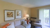 This single-floor space presents a thoughtfully arranged living area characterized by warm, golden-beige walls that create an inviting and comfortable ambiance. The textured ceiling adds a classic architectural detail overhead, complementing the overall design aesthetic of the room.

The living area is furnished with a plush, light-toned sofa accented by decorative pillows in neutral and terracotta hues, providing a cozy focal point for the space. A coordinating accent chair with patterned upholstery adds visual interest and additional seating. A dark wood side table positioned beside the sofa holds a classic table lamp, casting a warm, soft glow throughout the room.

Two framed art pieces displayed side by side on the wall contribute a curated, finished look to the space. A wall-mounted heating unit provides practical climate control while maintaining a clean profile along the lower wall. Natural light filters into the room through a window dressed with vertical blinds, offering privacy while still brightening the interior. The combination of warm tones, layered lighting, and carefully selected furnishings gives this space a relaxed yet polished character.