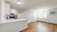 Located at 12000 Fairhill Rd in Cleveland, Ohio, this 2-bedroom, 1-bathroom unit offers a clean, modern aesthetic throughout its efficiently designed single-floor layout. The open-concept kitchen and living area form the heart of the home, featuring warm hardwood-style flooring, white shaker cabinetry, light stone countertops with a peninsula, and a full suite of stainless steel appliances including a refrigerator, range, microwave, and dishwasher. A matte black faucet adds a contemporary accent to the kitchen sink. The living area is bright and welcoming, with a large window fitted with horizontal blinds that allows ample natural light to fill the space. The primary bedroom is a comfortable, well-proportioned room with its own window, a wall-mounted air conditioning unit, and baseboard heating. The second bedroom features similar hardwood-style flooring in a cooler grey tone and a large window. A dedicated walk-in closet provides generous storage with built-in shelving and hanging rods. The bathroom is sleekly appointed with a white vanity featuring gold and black hardware, a framed mirror, and a glass-enclosed walk-in shower. Consistent light tones and clean finishes throughout create a bright, cohesive ambiance.