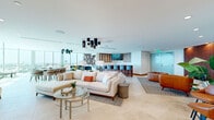Located at 795 Third Ave, this elegantly designed lounge space offers a refined, open-plan environment spread across a single floor. The space is anchored by dramatic floor-to-ceiling glass walls that flood the interior with abundant natural light and provide sweeping panoramic views of the surrounding cityscape. Large-format travertine-style tile flooring runs throughout, lending a cohesive and upscale aesthetic to the entire space.

The living area is thoughtfully organized into distinct functional zones. A plush, cream-toned sectional sofa paired with a glass-top coffee table forms the central lounge area, accented by a geometric patterned area rug. Adjacent seating vignettes feature rich cognac leather chairs and sofas, adding warmth and contrast. Striking cluster pendant light fixtures in matte black serve as bold focal points overhead.

A bar-height counter with rows of blue upholstered stools defines the bar and dining area, complemented by warm wood-toned cabinetry and a decorative tile backsplash. An open wood-grid shelving feature wall adds architectural interest and visual depth. Additional seating alcoves with casual chairs and small tables are positioned near the window walls, inviting relaxed conversation with city views as a backdrop. Indoor greenery is tastefully integrated throughout, enhancing the space's fresh, modern ambiance.