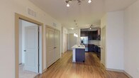 This 1-bedroom, 1-bathroom residence is designed with an open, functional layout that connects the entryway corridor seamlessly to the kitchen and living areas. The kitchen serves as the focal point of the space, featuring dark espresso cabinetry that contrasts elegantly with light stone countertops. A center island with a built-in sink provides additional prep space and doubles as a casual dining or gathering spot. Stainless steel appliances, including a French door refrigerator with an in-door water and ice dispenser, a dishwasher, and a microwave, complete the modern kitchen setup. Pendant lighting above the island and track lighting along the ceiling ensure the space is well-lit throughout. Warm hardwood-style flooring runs continuously through the main living areas, adding a cohesive and inviting feel to the interior. The hallway off the main corridor leads to additional rooms, with multiple doors suggesting well-organized closet and storage options. High ceilings contribute to an airy, open atmosphere throughout the unit, enhancing the sense of space. Overall, the unit offers a clean, contemporary aesthetic with practical design elements.
