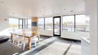 The Dwell Club Room is a thoughtfully designed communal gathering space that combines comfort, functionality, and style in a single open-plan layout. The room is bathed in natural light, thanks to an expansive array of black-framed windows that wrap around the dining and entertaining area, offering sweeping views of the surrounding neighborhood and outdoor terrace access through a full-height glass door.

The dining zone anchors the central space with a rustic wood farmhouse table paired with white modern chairs, set atop a patterned area rug that adds texture and warmth to the contemporary flooring beneath. A wall-mounted television is positioned on a natural wood accent panel, making this area equally suited for presentations or casual viewing.

Transitioning toward the lounge, a generously sized tufted black leather sectional sofa commands attention, offering ample seating for relaxed gatherings or socializing. A matching tufted ottoman completes the set, while framed local artwork adorns the crisp white walls, adding a sense of place and character to the space.

Adjacent to the main room, a sleek kitchenette features gloss white cabinetry, a stainless steel sink, a full-size refrigerator, and a microwave — providing all the essentials for hosting events or casual get-togethers. A window above the kitchenette maintains the bright, airy feel consistent throughout the room.