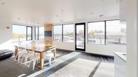 The Dwell Club Room is a thoughtfully designed communal gathering space that combines comfort, functionality, and style in a single open-plan layout. The room is bathed in natural light, thanks to an expansive array of black-framed windows that wrap around the dining and entertaining area, offering sweeping views of the surrounding neighborhood and outdoor terrace access through a full-height glass door.

The dining zone anchors the central space with a rustic wood farmhouse table paired with white modern chairs, set atop a patterned area rug that adds texture and warmth to the contemporary flooring beneath. A wall-mounted television is positioned on a natural wood accent panel, making this area equally suited for presentations or casual viewing.

Transitioning toward the lounge, a generously sized tufted black leather sectional sofa commands attention, offering ample seating for relaxed gatherings or socializing. A matching tufted ottoman completes the set, while framed local artwork adorns the crisp white walls, adding a sense of place and character to the space.

Adjacent to the main room, a sleek kitchenette features gloss white cabinetry, a stainless steel sink, a full-size refrigerator, and a microwave — providing all the essentials for hosting events or casual get-togethers. A window above the kitchenette maintains the bright, airy feel consistent throughout the room.