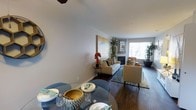 This 1-bedroom, 1-bathroom single-floor residence offers a thoughtfully designed layout that seamlessly connects the living and dining areas into one cohesive, inviting space. Rich hardwood flooring flows throughout, anchoring the warm and contemporary aesthetic. The dining area features a round glass table with stylish seating, positioned to take advantage of the open sightlines into the living room. The living room is a comfortable and well-lit space, highlighted by a classic fireplace that serves as a natural focal point, flanked by lush indoor plants that add a touch of greenery and life to the room. Large windows allow generous natural light to pour in, creating an airy and welcoming ambiance. Neutral-toned walls and carefully chosen furnishings — including a cushioned sofa and accent chairs with warm orange and blue accents — contribute to a balanced, modern interior. A framed piece of artwork above the sofa adds a decorative touch that ties the space together. The overall layout is functional and elegant, offering residents a comfortable and stylish environment suited for both everyday living and entertaining.