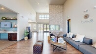 Located in Monument, Colorado, this single-floor leasing office for MarQ Apartments offers a well-appointed and professionally designed environment spanning a generous footprint. The welcoming lobby greets visitors with soaring double-height ceilings, warm wood-look flooring, and a floor-to-ceiling stacked stone accent wall flanking a sleek modern fireplace. A plush sectional sofa, organic decor accents, and abundant natural light from large windows create an inviting, comfortable atmosphere for prospective residents. An open transition leads into the kitchen and living area, where rich dark wood cabinetry is paired with granite-style countertops, floating wood shelves with greenery, and a full-size stainless steel refrigerator. A white breakfast bar with bar-top seating extends into the open space, offering a casual and functional gathering point. The adjacent office workspace is outfitted with built-in countertop desks, dark cabinetry, granite counters, and a wall-mounted display screen, all set against a calming blue accent wall. Carpeted flooring and recessed lighting lend a professional tone to this area. Private rooms accessible via charcoal-paneled doors offer additional functional space. The overall design balances modern aesthetics with warm, natural elements, reflecting a polished and welcoming professional setting.