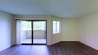 This single-floor space features a clean, open layout designed to maximize natural light and outdoor connectivity. The living area is finished with rich, dark hardwood-style flooring that contrasts attractively against the soft yellow-toned walls, creating a warm and inviting atmosphere. A textured ceiling adds subtle visual interest overhead.

A large sliding glass door serves as a focal point of the main living area, offering direct access to an outdoor balcony. The balcony overlooks mature, lush greenery, providing a sense of privacy and a tranquil natural backdrop. Horizontal blinds on the adjacent window allow for adjustable light control while maintaining the airy feel of the space.

A secondary window on the adjoining wall further enhances natural light penetration, ensuring the space feels open and well-lit throughout the day. The minimalist design of the interior allows for flexible furniture arrangement and personalization. White baseboards provide a crisp, clean finish along the perimeter of the room. Overall, the space presents a comfortable, light-filled environment with pleasant outdoor views and a straightforward, functional layout.