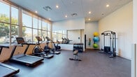 Located at 1311–1355 Decatur St in Denver, CO, this single-level fitness center offers a comprehensive range of exercise equipment within a clean, modern environment. The space is defined by a striking wall of large, grid-pane windows that span nearly the full length of one side, flooding the interior with natural light and offering open views of the surrounding outdoor greenery. The durable, dark rubber flooring throughout provides a practical and resilient surface suitable for a variety of workouts.

The cardio zone features multiple treadmills, elliptical trainers, and a stationary cycling bike arranged along the windowed wall, allowing users to exercise while enjoying the outdoor views. A large wall-mounted mirror runs along an adjacent wall, visually expanding the space and supporting functional training. The strength training area is well-appointed with a multi-tiered dumbbell rack offering a full range of free weights, an adjustable weight bench, a cable resistance machine, and a collection of wall-mounted medicine balls in multiple sizes. Recessed lighting in the drop ceiling provides consistent illumination throughout. The facility also includes accessible restroom facilities, clearly marked exits, and open floor space for stretching or floor exercises, contributing to a well-rounded and functional fitness environment.