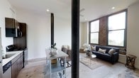 This single-floor space masterfully combines industrial character with contemporary design. The open-plan layout is anchored by striking exposed structural steel columns with riveted detailing, which serve as bold architectural focal points throughout the interior. Original exposed brick walls add warmth and texture, contrasting beautifully against crisp white walls and smooth polished concrete floors.

The living and dining areas flow seamlessly into one another, creating a cohesive and versatile entertaining space. Large windows with classic framing flood the interior with generous natural light, offering views of the surrounding greenery and enhancing the airy, open ambiance. Recessed lighting and ceiling fans complement the natural light, ensuring comfort throughout the day.

The dining area features a sleek stainless steel table with upholstered seating, while the living area is appointed with a contemporary dark sofa, a glass-top round coffee table, and a layered area rug that adds softness to the polished concrete underfoot. Carefully placed greenery and floral arrangements bring life and color to the space. Overall, the design strikes a refined balance between raw, industrial heritage and modern residential comfort.
