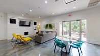 This single-floor space offers a thoughtfully designed open-plan layout that seamlessly integrates multiple functional zones within one expansive interior. Polished concrete floors flow throughout, lending an industrial-chic character that ties the entire space together cohesively.

The lounge area serves as a central gathering point, anchored by a large L-shaped sectional sofa in charcoal gray, accented with teal and sage throw pillows. Bold yellow accent chairs add a striking pop of color. A shiplap-clad feature wall houses a wall-mounted flat-screen television above a sleek fireplace, creating a strong focal point. Framed botanical artwork adds visual interest to the white walls, while recessed lighting keeps the atmosphere bright and inviting. Glass doors open directly to an outdoor balcony, flooding the space with natural light and offering views of surrounding greenery.

Adjacent to the lounge, a round dining table with transparent teal chairs provides a stylish seating area. A shuffleboard table and additional dining seating extend the space's entertainment capabilities further.

The kitchen is efficiently appointed with stainless steel appliances, including a dishwasher, electric range, and microwave. Dark cabinetry contrasts cleanly against white subway tile backsplash and countertops, while floating shelves and a large peninsula with bar seating add both storage and casual dining functionality. A pendant light anchors the kitchen's aesthetic with an industrial touch.