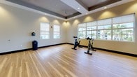 This single-floor fitness studio is thoughtfully designed to accommodate a variety of exercise activities, from cycling classes to yoga and group fitness sessions. The space is anchored by smooth, light-toned hardwood flooring that extends throughout, providing a clean and durable surface well-suited for physical activity.

The studio features a coffered ceiling with recessed lighting and ceiling fans, creating a comfortable, evenly lit environment. Large windows with roller shades line the walls, allowing generous natural light to filter in while offering views of the surrounding outdoor landscape, including palm trees and neighboring buildings. The windows give the space a bright, open feel that enhances the workout atmosphere.

A full wall of floor-to-ceiling mirrors runs along one side of the room, expanding the sense of space and allowing users to monitor their form during exercise. Several spin bikes are positioned near the windowed walls, taking advantage of the natural light and views. A large wall-mounted flat-screen display is centrally installed on the main wall, suitable for instructor-led video workouts or class programming.

Glass-paneled doors connect to adjacent areas, and the neutral warm-toned walls contribute to the studio's clean, modern aesthetic overall.