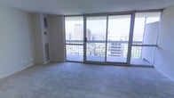 This high-rise living space is defined by its striking wall of floor-to-ceiling sliding glass doors and windows, which flood the interior with abundant natural light and frame a dramatic panoramic view of the surrounding urban skyline. The living area features wall-to-wall carpeting in a neutral tone, providing a clean and versatile foundation for furnishing. A sliding glass door opens directly onto a private balcony enclosed by a metal railing, offering an outdoor extension of the living space with elevated city views. The corner window configuration maximizes light exposure from multiple angles, enhancing the sense of openness throughout the room. A wall-mounted HVAC vent is positioned near the floor, ensuring climate control along the exterior glass wall. The smooth, light-toned walls and textured ceiling complement the bright, airy atmosphere created by the expansive glazing. The overall ambiance is bright and urban, with the elevated vantage point providing a compelling connection to the cityscape below, making the views a central architectural feature of the space.
