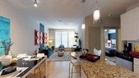 This single-floor residence offers a well-designed open-concept layout that seamlessly connects the kitchen, dining, and living areas. The kitchen is outfitted with stainless steel appliances, white shaker-style cabinetry, a white subway tile backsplash, and a granite-topped island with bar seating for two. Elegant pendant lighting with decorative glass fixtures hangs above the island, complementing the track lighting overhead. Hardwood-style flooring runs throughout the main living area, adding warmth to the space.

The living room is a visual standout, anchored by a bold navy blue accent wall adorned with two striking red-framed art panels featuring circular wood details. A teal velvet sofa and patterned area rug complete the contemporary aesthetic. A separate dining area with a glass-top table and white chairs sits adjacent to the living space.

The bedroom is bright and spacious, featuring large windows that fill the room with natural light, a ceiling fan, carpeted flooring, and a dramatic horizontal navy accent stripe along the wall. It connects to a generously sized walk-in closet with multi-level shelving and hanging rods. The bathroom includes a full soaking tub with tile surround. A dedicated laundry area with a full-size washer and dryer adds everyday convenience.