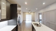 This modern single-floor space in Plano offers a clean, open-concept layout that seamlessly connects the kitchen and living areas. The kitchen is well-appointed with dark espresso cabinetry that provides a striking contrast against the white quartz countertops. A stainless steel undermount sink sits along the kitchen island, complemented by a stainless steel French door refrigerator and recessed lighting that illuminates the space effectively. The light wood-toned flooring runs consistently throughout, contributing to a warm and cohesive aesthetic. The living area extends naturally from the kitchen, featuring a ceiling fan with integrated lighting and recessed can lights, creating a well-lit and comfortable environment. A large window fitted with a white roller shade allows for controlled natural light, brightening the space while maintaining privacy. Neutral, warm-toned walls keep the interior feeling airy and versatile. A doorway visible on the right side of the space hints at additional rooms, suggesting a functional and thoughtfully organized floor plan. Overall, the space presents a contemporary design with quality finishes suited for modern living.