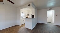 This single-floor residence offers a clean, modern aesthetic throughout, anchored by rich, dark hardwood-style flooring that flows seamlessly across all living areas. The open-concept layout connects the living room to the kitchen, creating a spacious and functional living environment. The living room is generously sized, featuring a ceiling fan, bright white walls, and large windows with vertical blinds that invite natural light. A front entry door flanked by two windows adds additional brightness to the space.

The kitchen is framed by a peninsula-style breakfast bar that defines the transition from the living area. It features crisp white cabinetry with matte black hardware, light-toned countertops, a deep basin sink, and a full suite of stainless steel appliances including a range, over-the-range microwave, and refrigerator. Track lighting illuminates the kitchen workspace effectively.

A dedicated bedroom area is accessible from the main living space, offering a ceiling fan and a large sliding glass door with vertical blinds, providing access to natural light and outdoor views. The bathroom is finished with wood-look flooring, a full bathtub and shower combination with a rain-style showerhead, and matte black fixtures throughout, maintaining a consistent modern design language across the entire residence.