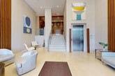 Located at 1 Ocean Drive in Brooklyn, NY 11224, this sophisticated building lobby makes a strong first impression with its refined architectural design and upscale finishes. Floor-to-ceiling glass entry doors flood the space with natural light, offering clear sightlines to the surrounding streetscape and creating an open, welcoming atmosphere from the moment of arrival.

Inside, the lobby is anchored by a stunning reception desk that pairs a live-edge wood top with a dramatic marble base featuring bold veining — a centerpiece that seamlessly blends natural materials with contemporary design. Tall, floor-to-ceiling slatted wood accent walls line multiple areas of the lobby, adding warmth, texture, and visual depth throughout the space.

The expansive layout is finished with large-format cream tile flooring that enhances the sense of scale and brightness. Multiple seating areas are thoughtfully appointed with modern white and gray upholstered chairs and sofas, offering comfortable gathering spots for residents and visitors. Contemporary pendant lighting and recessed ceiling fixtures provide layered illumination, while curated artwork on the walls adds a gallery-like quality to the environment.

A grand staircase with polished marble treads and sleek metal railings serves as a focal architectural element, complemented by a stainless steel elevator nearby. The overall ambiance is calm, polished, and modern — befitting a premier address along Brooklyn's waterfront.