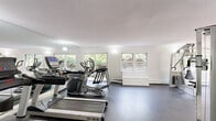 Located at 495 Laurel Hill Rd, this dedicated fitness facility offers a comprehensive workout environment across a single floor. The main exercise room is the centerpiece of the space, featuring durable rubber flooring well-suited for high-intensity training. Large windows along multiple walls flood the room with natural light and provide views of the surrounding lush greenery, creating an inviting and energizing atmosphere. Full-length mirrors are strategically placed to enhance the sense of openness and support proper form during exercise. The room is outfitted with a wide array of professional-grade equipment, including treadmills, elliptical machines, a cable machine system by Cybex, and additional strength-training apparatus, accommodating both cardio and resistance training needs. Sliding glass doors on one side provide direct access to the outdoors, further connecting the interior to the natural surroundings. The facility also includes a well-appointed bathroom finished with wood-look tile flooring, white tile walls, a glass-enclosed shower/tub combination, and a clean white vanity — ideal for freshening up post-workout. A hallway connects the key areas of the space, ensuring smooth and functional circulation throughout.