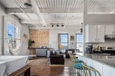 This character-rich loft-style residence blends industrial heritage with modern comfort across a single, expansive floor. Rich reclaimed hardwood floors run throughout, complemented by soaring ceilings with exposed wooden beams, structural columns, and industrial ductwork that speak to the building's storied past. The open-concept kitchen and living area serves as the heart of the home, featuring a large granite-topped island with bar seating, white cabinetry, stainless steel appliances, and pendant lighting overhead. The living room is anchored by large multi-pane windows that flood the space with natural light and city views, an exposed brick accent wall, and a sleek wall-mounted electric fireplace set against a reclaimed wood surround. A long gallery-style hallway lined with framed artwork leads to the private quarters. The generously sized primary bedroom showcases another stunning exposed brick wall, tall windows, and soaring ceilings with an eye-catching chandelier. The bathroom features a granite-topped vanity with a round mirror and a full bathtub with shower. A dedicated laundry area with a full-size washer and dryer adds everyday convenience. This space offers a distinctive urban loft aesthetic with warmth and character throughout.