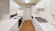 This single-floor residence at 12715 Vinings Creek Dr in Matthews, NC offers a practical and well-organized layout. The galley-style kitchen is outfitted with white cabinetry featuring brass hardware, white appliances including a coil-burner range and refrigerator, a stainless steel double sink, and wood-look flooring. A bi-fold closet door at the end of the kitchen provides additional storage. The kitchen opens toward the living area via a peninsula with a breakfast bar, creating a connected flow between the two spaces. The living area features neutral carpet, a ceiling fan, and access to an outdoor area through a sliding glass door, allowing natural light to fill the room. Bedrooms are carpeted and finished with neutral tones; one includes a large window with views of surrounding greenery, while another features its own sliding glass door access. Two full bathrooms each include a tiled tub-shower combination, a vanity with a large mirror, and bright overhead lighting. Textured ceilings are present throughout, and the overall color palette of whites and soft neutrals gives the space a clean, functional character suitable for everyday living.