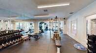 This fitness center offers a comprehensive workout facility spread across a generously sized single-floor layout. The space is divided into distinct functional zones, each designed to support a full range of exercise activities. The cardio area features an impressive vaulted ceiling with exposed wood beams and tongue-and-groove paneling, ceiling fans, and large windows that frame views of an outdoor pool and surrounding greenery, filling the space with natural light. Treadmills, an elliptical, and a stair climber are positioned to take full advantage of the scenic outlook. The strength training zone is equipped with a multi-station cable machine, adjustable benches, and a well-stocked dumbbell rack ranging across multiple weight increments. Floor-to-ceiling mirrors line one wall, visually expanding the space and providing form-checking visibility throughout. Durable rubber-tile flooring covers the entire facility, offering both safety and functionality. Overhead linear lighting ensures consistent illumination throughout. A separate utility area includes louvered closet doors, a mounted water fountain, and posted fitness center guidelines. The overall design balances practical amenity with an inviting, airy ambiance suitable for a community fitness facility.