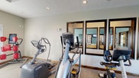 This single-floor community clubhouse offers a well-appointed gathering space designed for comfort and functionality. The main lounge area makes an immediate impression with soaring ceilings and an elegant chandelier casting warm light over neutral-toned seating, including a tufted cream sofa, wingback chairs, and a casual dining area with a wooden table. A dramatic floor-to-ceiling stacked stone fireplace with a rustic wood mantel serves as the focal point of the room, adding warmth and character to the space. Dark-framed French doors and large transom windows flood the interior with natural light and provide access to an outdoor area, seamlessly connecting the indoor and outdoor environments. The lounge transitions through glass-paned interior doors into additional rooms, including a teal-accented space visible beyond, suggesting flexible use for meetings or social gatherings. The fitness center is practical and well-equipped, featuring multiple Octane brand elliptical trainers and a stationary bike arranged on carpeted flooring beneath recessed lighting. Large dark-framed mirrors line the fitness room wall, enhancing the sense of space. Throughout the clubhouse, consistent dark wood trim, neutral walls, and textured ceilings tie the design together into a cohesive, welcoming aesthetic.