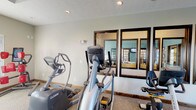 This single-floor community clubhouse offers a well-appointed gathering space designed for comfort and functionality. The main lounge area makes an immediate impression with soaring ceilings and an elegant chandelier casting warm light over neutral-toned seating, including a tufted cream sofa, wingback chairs, and a casual dining area with a wooden table. A dramatic floor-to-ceiling stacked stone fireplace with a rustic wood mantel serves as the focal point of the room, adding warmth and character to the space. Dark-framed French doors and large transom windows flood the interior with natural light and provide access to an outdoor area, seamlessly connecting the indoor and outdoor environments. The lounge transitions through glass-paned interior doors into additional rooms, including a teal-accented space visible beyond, suggesting flexible use for meetings or social gatherings. The fitness center is practical and well-equipped, featuring multiple Octane brand elliptical trainers and a stationary bike arranged on carpeted flooring beneath recessed lighting. Large dark-framed mirrors line the fitness room wall, enhancing the sense of space. Throughout the clubhouse, consistent dark wood trim, neutral walls, and textured ceilings tie the design together into a cohesive, welcoming aesthetic.