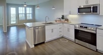 This 640-square-foot, one-bedroom, two-bathroom apartment at 2300 Hooks St, Unit 5105 in Clermont, FL offers a well-designed single-floor layout that maximizes space and functionality. The open-concept kitchen and living area serves as the heart of the unit, featuring wood-look plank flooring throughout, neutral warm-toned walls, and abundant natural light from multiple windows fitted with horizontal blinds. The kitchen is outfitted with white shaker-style cabinetry, a white subway tile backsplash, stainless steel appliances including a refrigerator, range, and microwave, and a large peninsula island with pendant lighting — ideal for dining or entertaining. The living area extends to a private exterior door with additional windows, providing outdoor access and views of the surrounding greenery. The 140-square-foot primary bedroom includes a ceiling fan with light, dual windows, and direct access to the en-suite bathroom. The primary bath features a single-sink vanity with gray countertops, white cabinetry, a soaking tub with shower, and a built-in tile niche. A spacious walk-in closet with custom shelving and dual hanging rods provides ample storage. A second full bathroom serves guests. Ceiling fans with lights are present in the main living areas and bedroom.