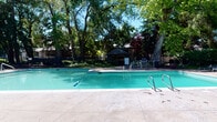 This outdoor amenity area centers around a generously sized swimming pool with clear turquoise water, accessible via stainless steel entry rails on both sides. The pool is surrounded by a broad, light-toned concrete deck that provides ample space for lounging and recreation. Mature deciduous trees, including large shade trees with full canopies, frame the perimeter, creating a naturally shaded and visually appealing environment during warm months.

At the far end of the pool, a covered gazebo with a dark octagonal roof serves as a shaded gathering spot, accompanied by patio seating and tables nearby. The pool area is fully enclosed by a black metal security fence with a latched gate entrance, ensuring controlled access. Safety equipment, including a life ring, is mounted near the covered utility corridor, which also stores pool maintenance tools and equipment.

Adjacent to the pool area is an enclosed fitness facility, visible through large windows. The gym features ceiling fans, recessed lighting, rubber flooring, and exercise equipment, offering an air-conditioned workout space directly accessible from the pool deck. The covered corridor connecting these spaces features a concrete floor and panel siding. Together, these amenities form a well-equipped, self-contained recreational complex.