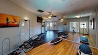 Located at 4802 N 12th St in Phoenix, Arizona, this dedicated fitness center offers a thoughtfully organized, single-floor workout facility designed to accommodate a wide range of exercise routines. The space is divided into two distinct workout zones, each with its own character and function.

The main workout area features durable rubber flooring and is lined with large floor-to-ceiling mirrors that amplify the sense of space and allow users to monitor their form. This section is equipped with cardio machines including treadmills, an elliptical, and a multi-station cable weight machine, alongside adjustable benches and a wall-mounted flat-screen television. Sconce lighting and a textured ceiling complete the utilitarian yet functional aesthetic.

A secondary room, finished with warm hardwood flooring, provides a more open, versatile training environment. This area includes a spin bike, stand-up paddleboard balance trainers, and a functional rack system, with motivational workout posters adorning the grey-painted walls. A ceiling fan with an integrated light fixture keeps the space comfortable, and a windowed door allows natural light to fill the room, creating a bright and welcoming atmosphere.

Together, the two areas form a comprehensive fitness facility well-suited for both cardio conditioning and strength training, making it a valuable amenity for the surrounding community.