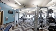 This fitness center offers a comprehensive workout environment designed to accommodate a variety of exercise routines. The space is thoughtfully laid out, with dedicated areas for both strength training and cardiovascular exercise. A row of treadmills and cardio machines lines the perimeter near large windows, allowing natural light to fill the room while offering views of the surrounding greenery outdoors.

The strength training area is outfitted with cable machines, weight racks, and benches, providing a full range of resistance equipment for users of all fitness levels. A wall-mounted television adds a functional amenity for those looking to stay entertained during their workout.

The interior design strikes a balance between functionality and style. The flooring features a bold chevron pattern that adds visual interest and clearly delineates the space. Light blue accent walls complement the neutral tones of the ceiling and equipment, creating an energizing yet comfortable atmosphere. Ceiling fans are evenly distributed throughout, ensuring proper air circulation during intense workouts.

Ample mirror panels are strategically placed to support proper form and visually expand the space. The combination of bright, even lighting from overhead fixtures and natural light from the large windows ensures a well-lit environment throughout the day.