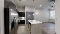 Located on Westheimer Road in Houston's prestigious Galleria corridor, this well-appointed one-bedroom, one-bathroom unit offers a thoughtfully designed layout across a single floor. The open-concept kitchen serves as the heart of the home, featuring a large center island with a built-in sink, crisp white countertops, rich dark cabinetry, a glass tile backsplash, and a full suite of stainless steel appliances including a gas range, microwave, and French door refrigerator. Pendant lighting above the island adds a contemporary touch, while stylish wood-look flooring flows consistently throughout the space. The living area benefits from natural light through large windows and includes a ceiling fan, with a private entrance door adding convenience. The spacious bedroom offers generous dimensions, a window with natural light, soaring ceilings, and direct access to a walk-in closet outfitted with multi-tier wire shelving for organized storage. The bathroom is elegantly finished with large-format tile flooring, a dual-sink vanity topped with granite countertops, dark wood cabinetry, a soaking tub, and a tall wood-paneled storage cabinet. Neutral gray walls and consistent dark-toned interior doors unify the modern aesthetic throughout the unit.