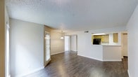 This single-floor residence offers an open and airy layout that seamlessly connects the main living, dining, and kitchen areas into one cohesive space. Rich, dark hardwood-style flooring runs throughout, providing a warm contrast to the neutral, light-toned walls and white trim that define the interior. The living and dining areas flow naturally into one another, with a pendant light fixture marking the dining space and adding a touch of understated elegance. Natural light filters in through a window dressed with classic horizontal blinds, brightening the open floor plan. A textured popcorn ceiling adds a traditional character to the space. The kitchen is accessible via a raised pass-through counter that overlooks the main living area, offering an open connection ideal for entertaining. The kitchen features warm wood-toned cabinetry and includes built-in appliances. A central hallway visible from the main area leads to the remaining rooms of the residence, suggesting a well-organized and functional floor plan. Overall, the space presents a clean, neutral palette that provides a versatile foundation for a variety of interior styles.